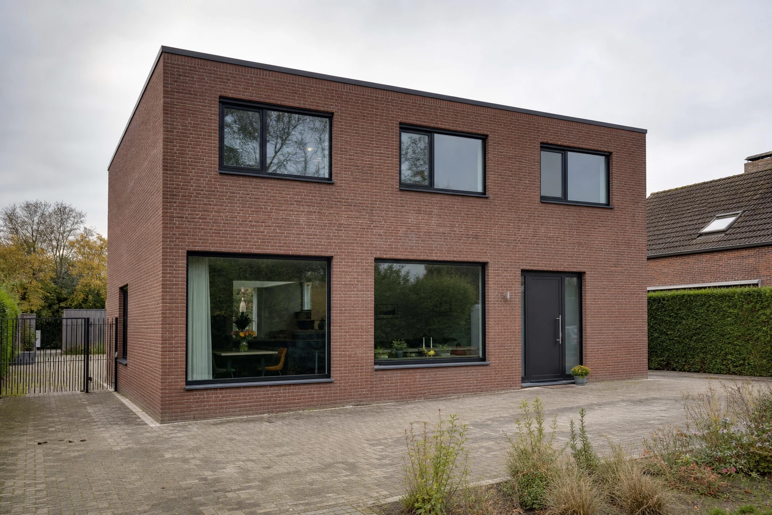 A flat-roofed house in red-brown brick with black window frames, a dark front door and a block-paved driveway